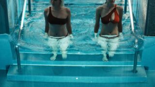 Alexandra Daddario and Julia Goldani Telles stepping into an indoor swimming pool together, both girls wearing swimsuits with Alexandra’s a bit more revealing with some cleavage in view