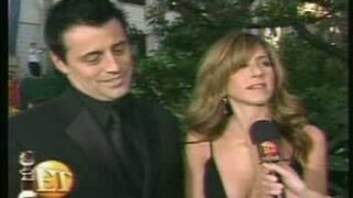 Jennifer Aniston wearing a black dress slit down to her stomach with a black strap across it and showing off some impressive cleavage as she interviews with a guy from Entertainment Tonight