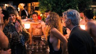 Margot Robbie wearing a pair of overalls with no top on underneath, showing plenty of side boob as she talks to some people at a party and then dances with Li Jun Li, who leans in to almost kiss her