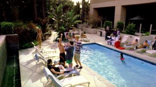 Vanessa Angel showing off her body in a bikini as she walks along a pool and then behind some guys as they’re getting ready to pose for a photo